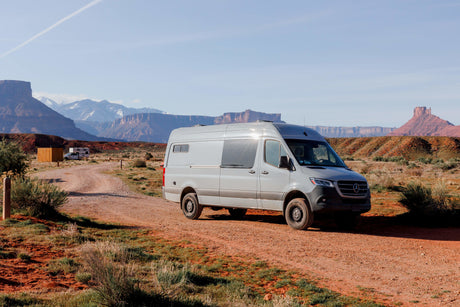 Grey Sprinter RV rental on a desert dirt road, with stunning red rock buttes and snow-capped mountains in view.
