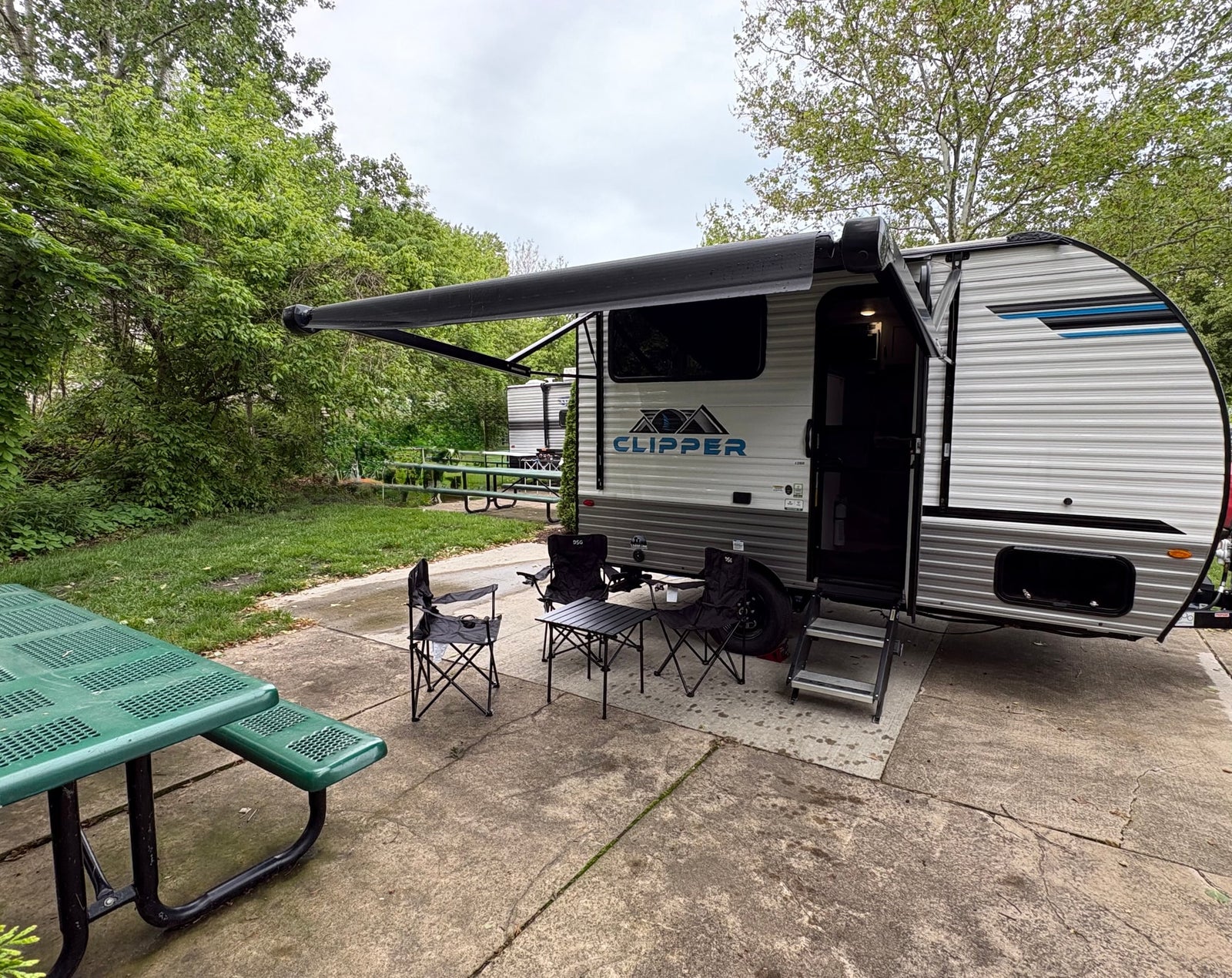 A modern RV rental travel trailer is set up at a peaceful campsite with an extended awning, chairs, and a picnic table.