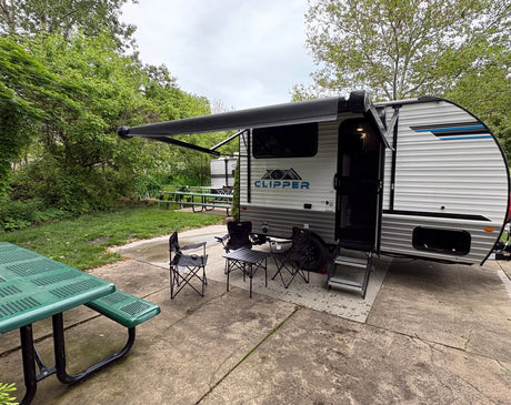 A modern RV rental travel trailer is set up at a peaceful campsite with an extended awning, chairs, and a picnic table.