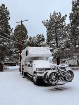 A snow-covered RV rental and bikes parked at a winter campsite, surrounded by frosty pine trees under a cloudy sky.