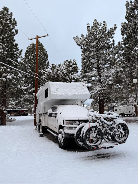 A snow-covered RV rental and bikes parked at a winter campsite, surrounded by frosty pine trees under a cloudy sky.