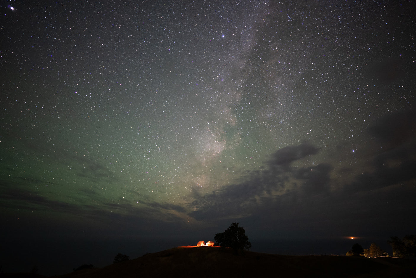 Milky Way and green airglow illuminate an RV rental, car, and tree on a hill, creating a stunning starry night camp scene.