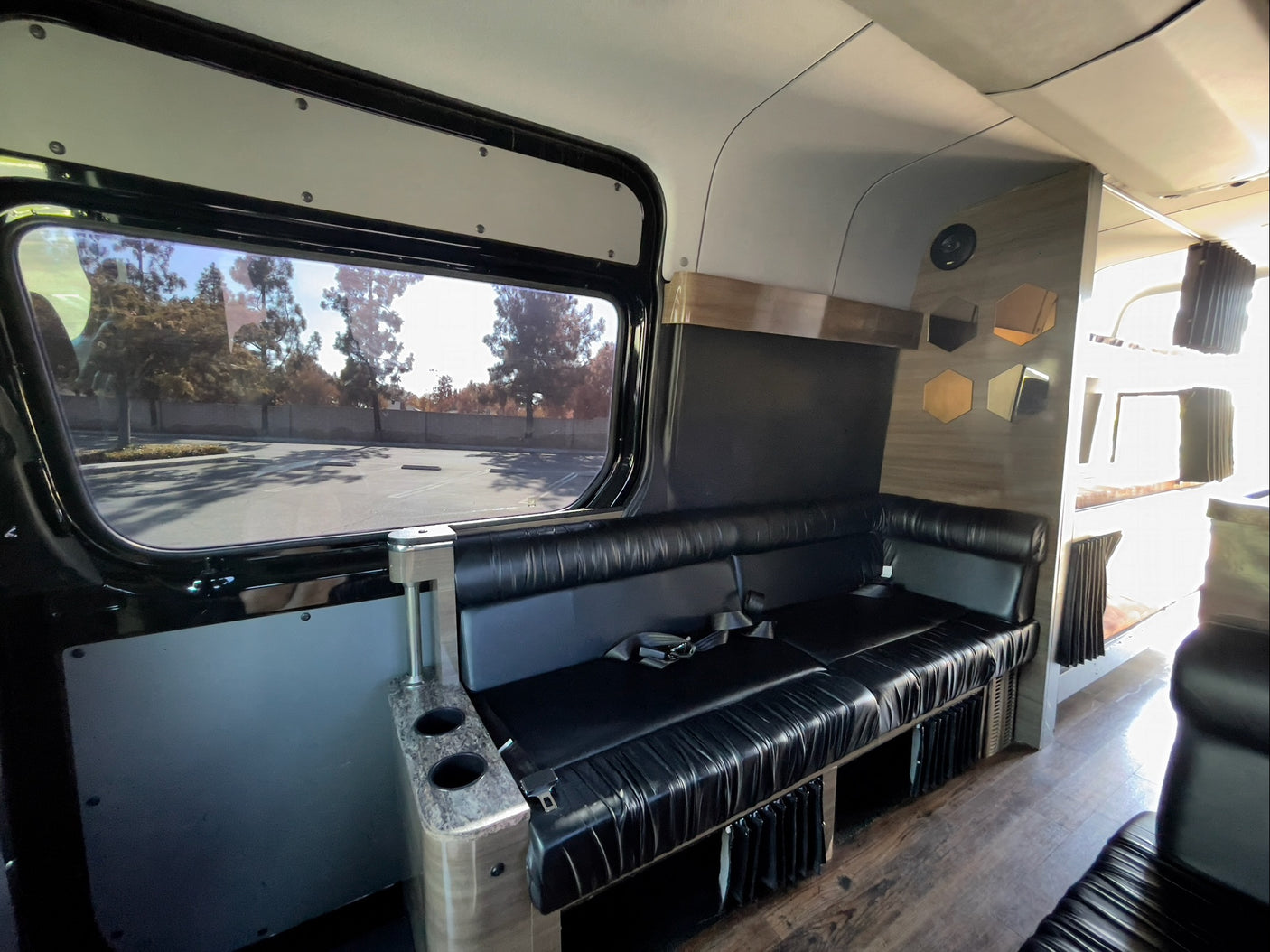 Modern RV rental interior: black leather bench seating, large window overlooking trees, wood floor, and wall decor.