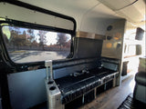Modern RV rental interior: black leather bench seating, large window overlooking trees, wood floor, and wall decor.