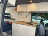 Modern RV rental interior: light wood kitchen counter with sink, white cabinets, shiplap walls. Bed and colorful decor for mobile living.