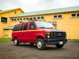 A vibrant red Ford E-Series camper van, an RV rental with Dakine racks, parked by a sunny yellow Hawaiian building.