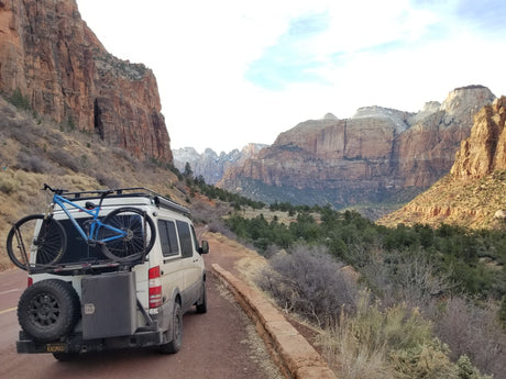 White camper van with bike on back, an ideal RV rental, on a dirt road in a stunning mountainous desert landscape.