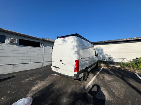 White Mercedes Sprinter van, ideal for RV rental and travel. Features a roof rack, parked under a clear blue sky.