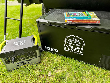 Preparing for an Alaska RV rental adventure with an Iceco cooler, Goal Zero power station, and travel guides on grass.