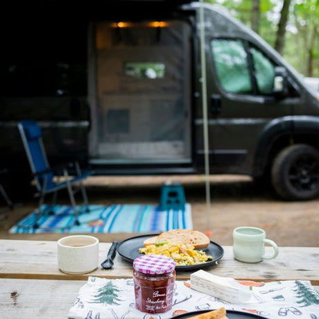 Morning spread: breakfast on a picnic table next to a parked rv rental van, with camping chair and mat outside.