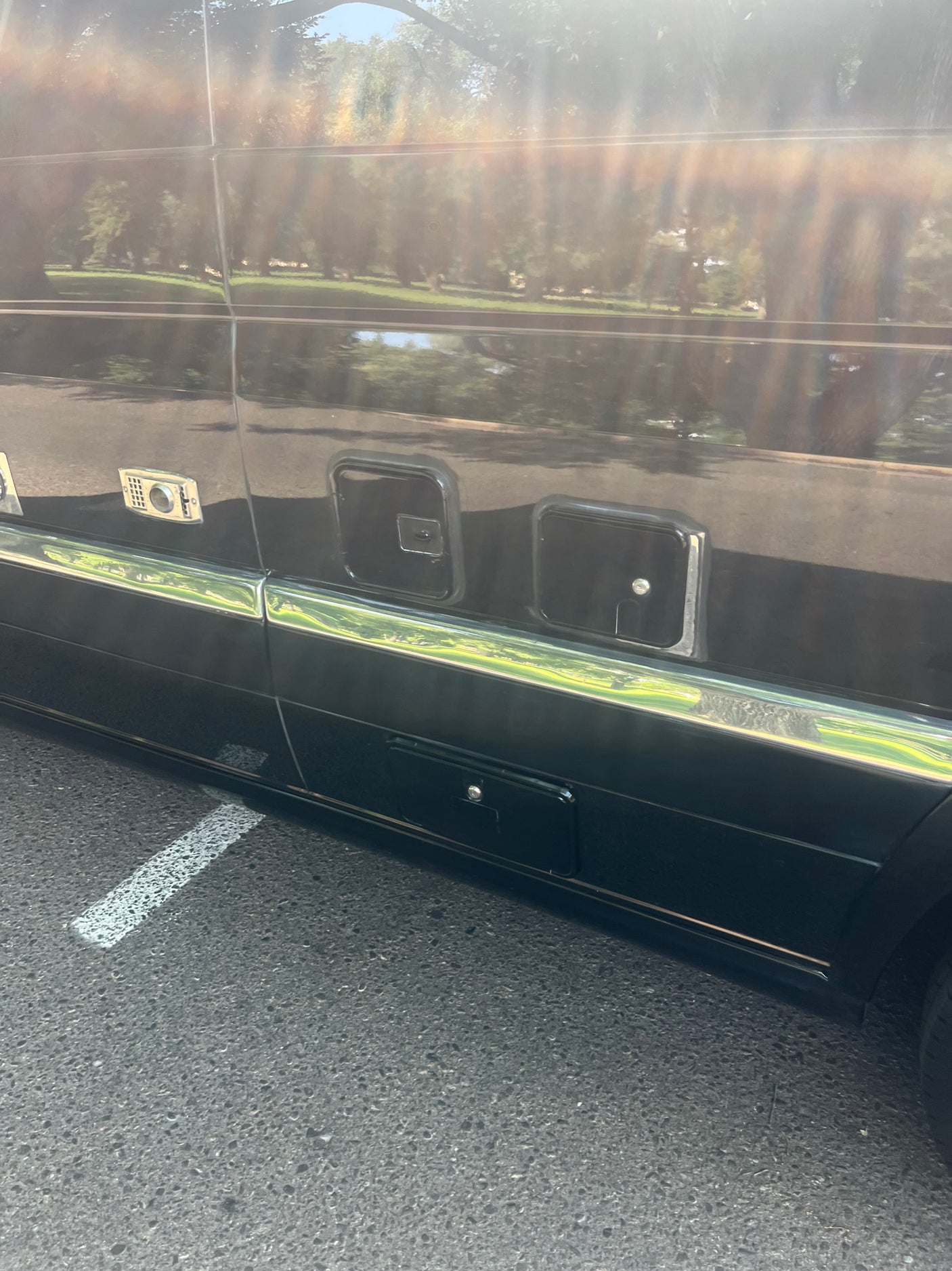 Side view of a dark RV rental, showing external compartments, a chrome strip, and reflections of trees.