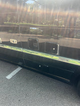 Side view of a dark RV rental, showing external compartments, a chrome strip, and reflections of trees.