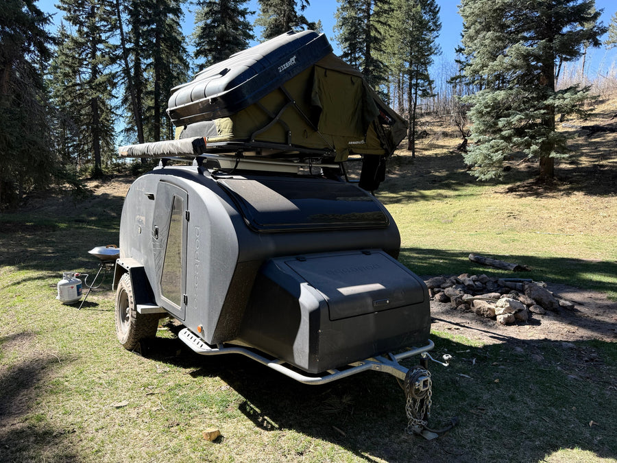 This off-road teardrop camper with a rooftop tent deployed in a treed campsite is perfect for an RV rental adventure.