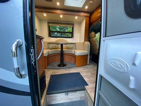 Inside a modern rv rental: a well-lit dinette with U-shaped seating, wood flooring, and a view to nature from the window.