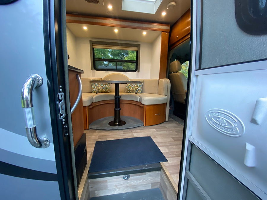 Inside a modern rv rental: a well-lit dinette with U-shaped seating, wood flooring, and a view to nature from the window.