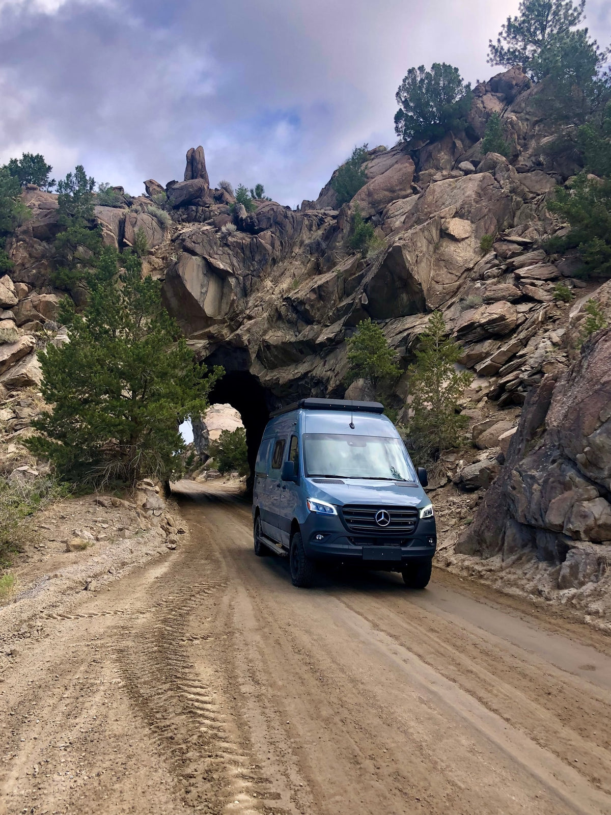 Blue Mercedes-Benz Sprinter RV rental van exits a rugged rock tunnel on a dirt road.