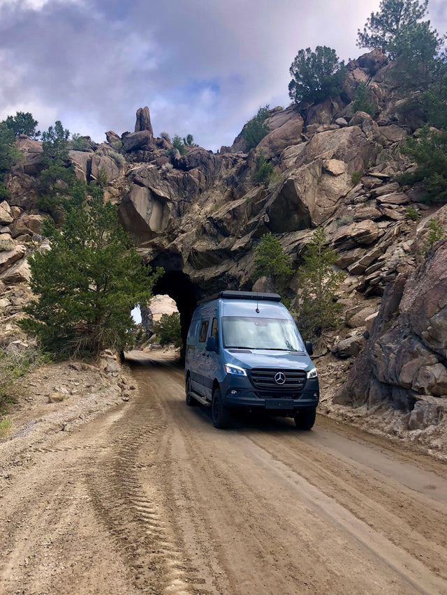 Blue Mercedes-Benz Sprinter RV rental van exits a rugged rock tunnel on a dirt road.