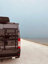 Gray Thor RV rental with a Thule rack parked on a dirt road by a calm bay under an overcast sky.