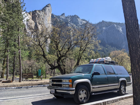 Dark green SUV with roof cargo (like an **RV rental**) against Yosemite's majestic mountains & waterfall for park exploration.