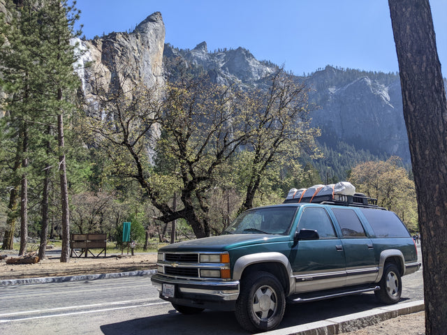 Dark green SUV with roof cargo (like an **RV rental**) against Yosemite's majestic mountains & waterfall for park exploration.
