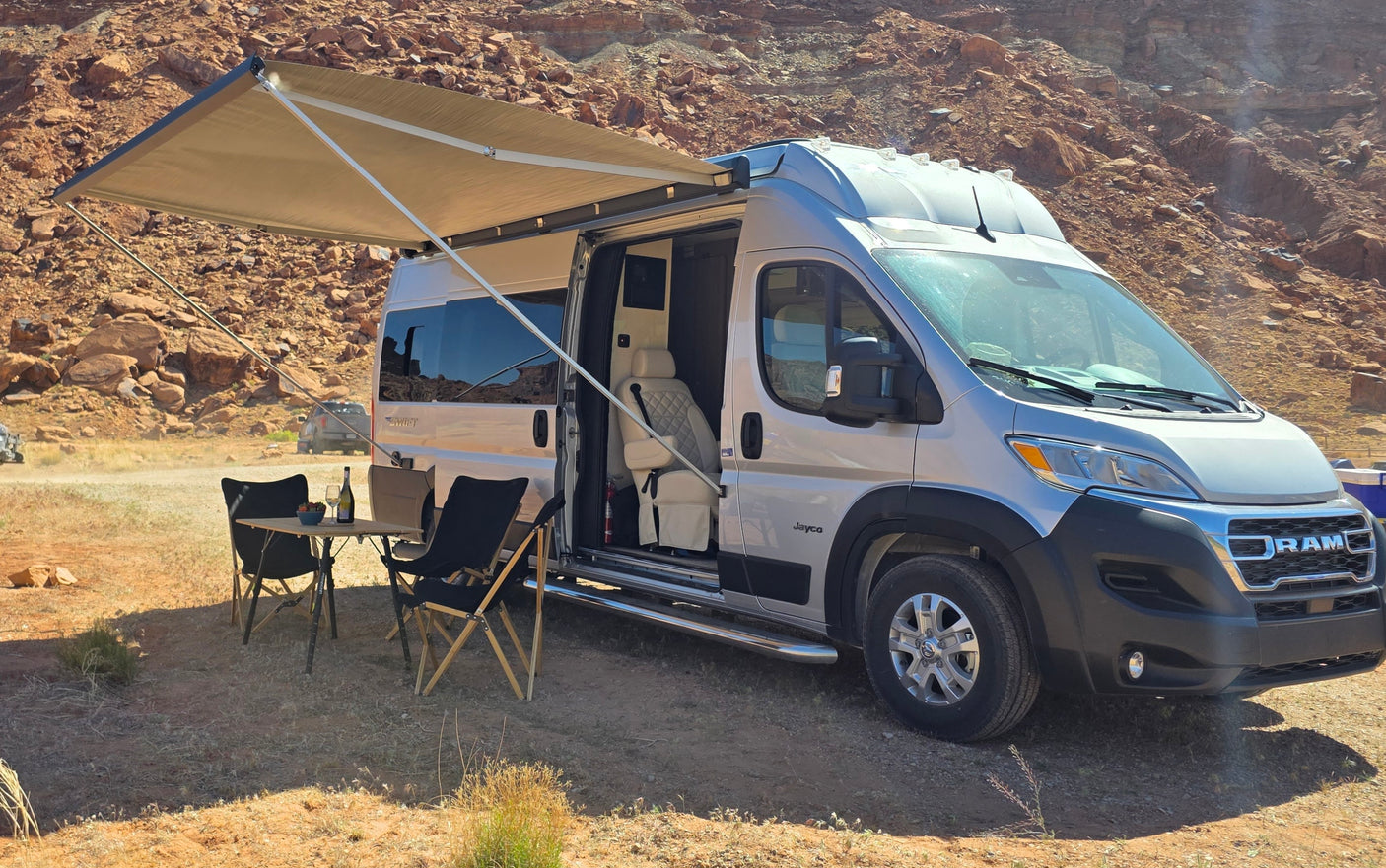 Desert boondocking with this RV rental. Camper van with open awning, chairs, and table set up in a rocky landscape.