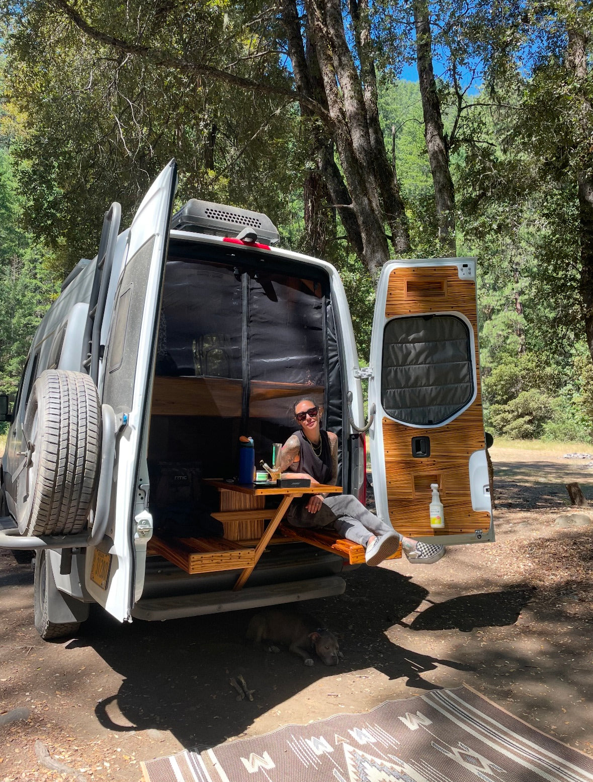 Relaxing in an RV rental camper van. A woman sits in the open back, dog rests below in a sunny forest setting.
