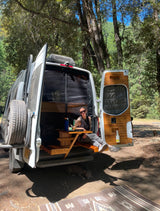 Relaxing in an RV rental camper van. A woman sits in the open back, dog rests below in a sunny forest setting.