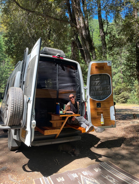 Relaxing in an RV rental camper van. A woman sits in the open back, dog rests below in a sunny forest setting.
