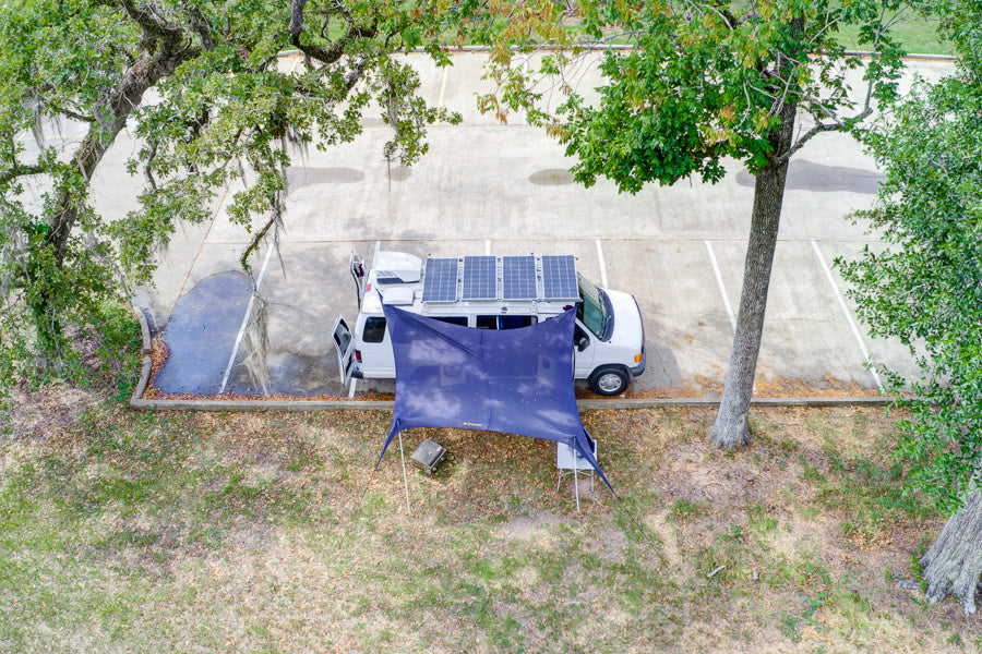 Overhead view of a white camper van, like an rv rental, with rooftop solar panels and a blue tarp awning set up on grass next to a parking lot.