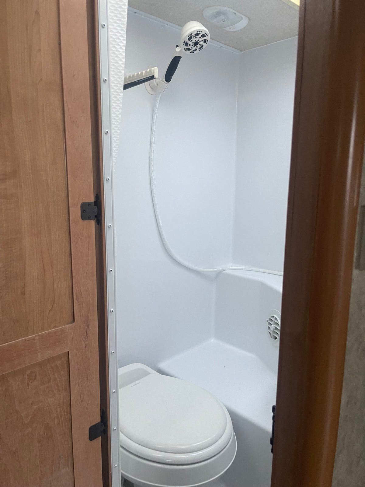 RV rental: A clean, compact white bathroom with a toilet and shower head on a hose. Wood trim frames the entry. — photo 7