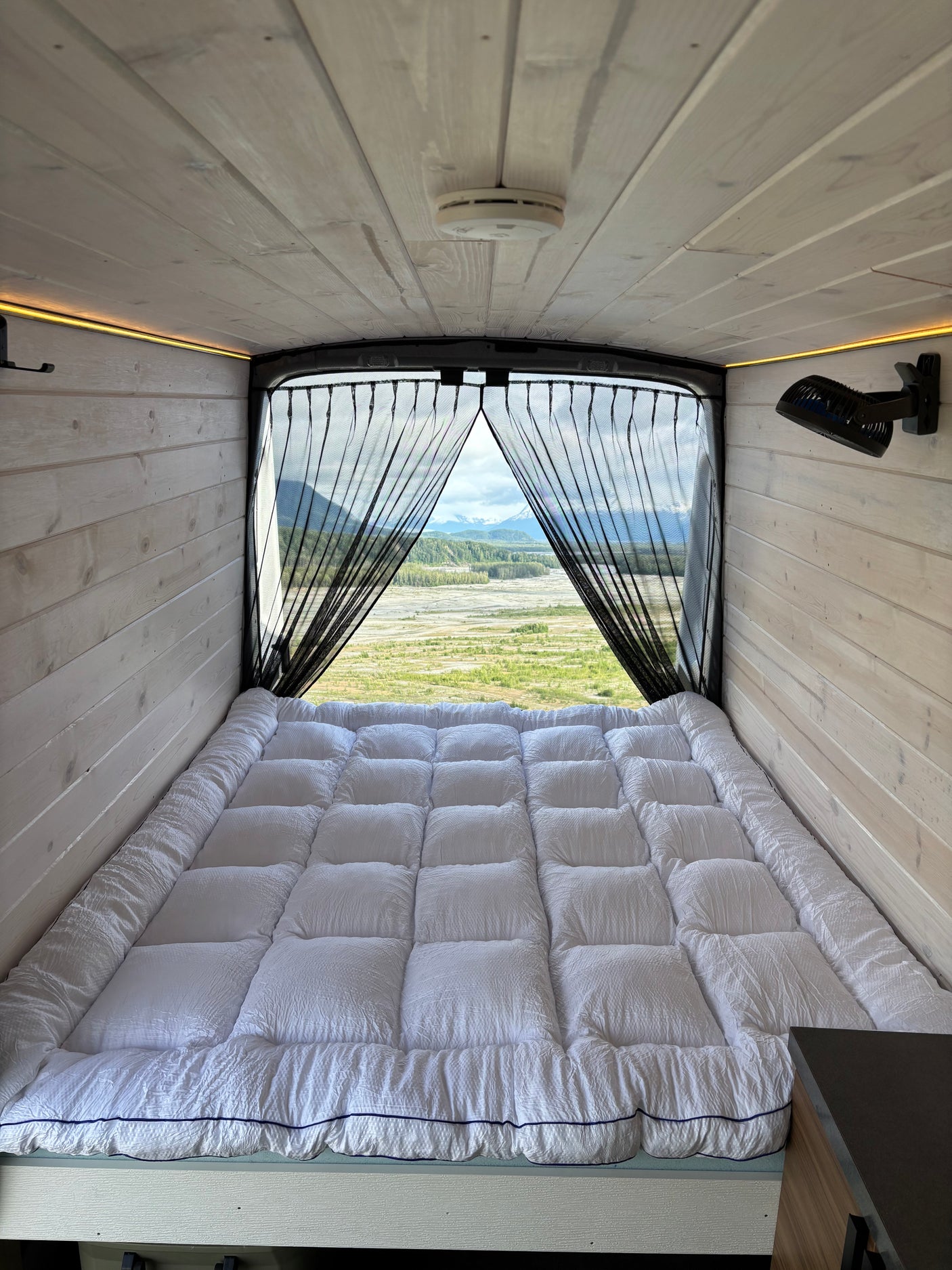Serene RV rental interior: cozy bed, warm wood paneling, and a breathtaking mountain and river view through the screen.