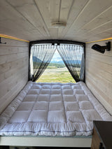 Serene RV rental interior: cozy bed, warm wood paneling, and a breathtaking mountain and river view through the screen.