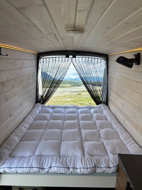 Serene RV rental interior: cozy bed, warm wood paneling, and a breathtaking mountain and river view through the screen.
