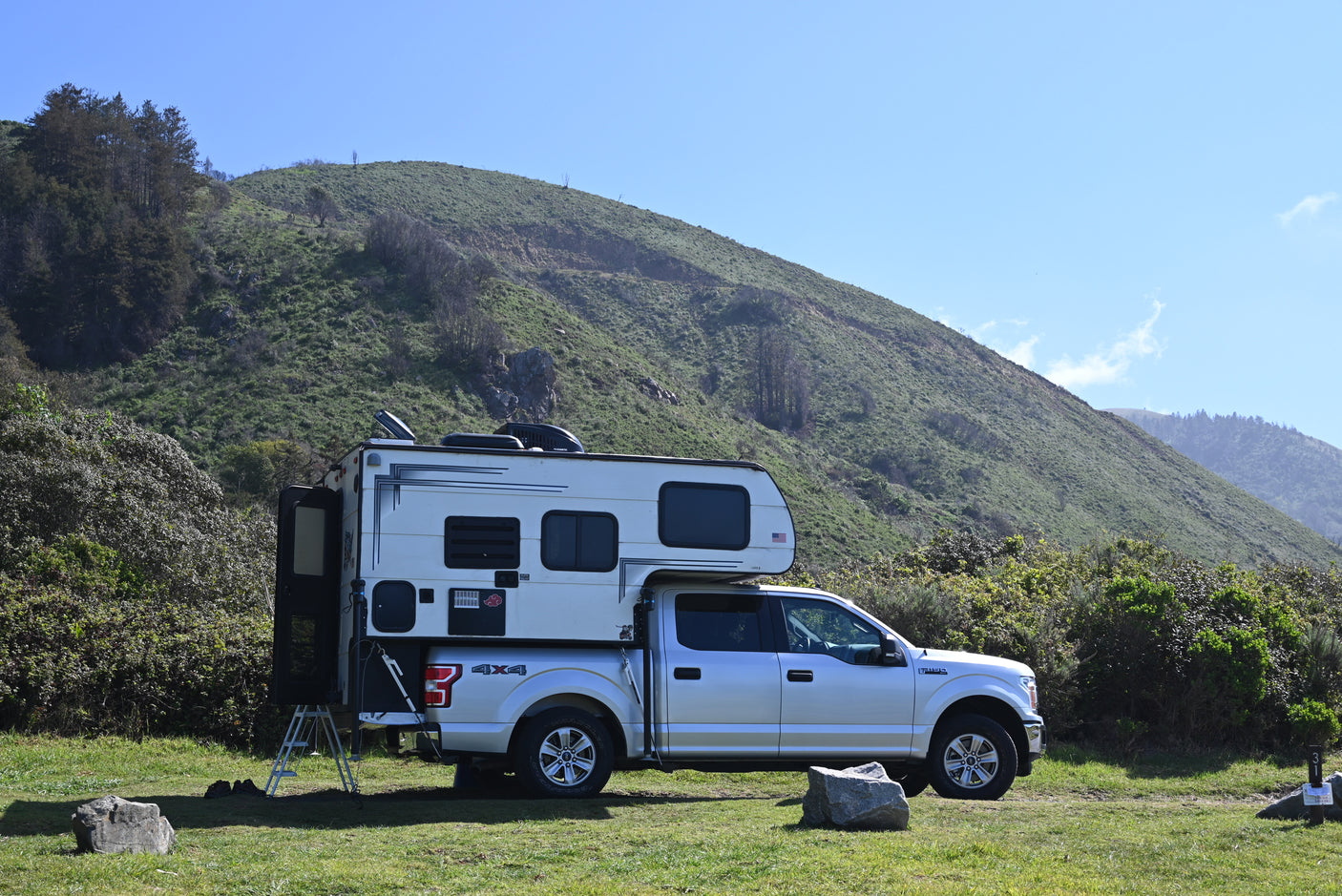 An RV rental truck camper on a silver pickup, parked on green grass with a large hillside behind it, ideal for camping.
