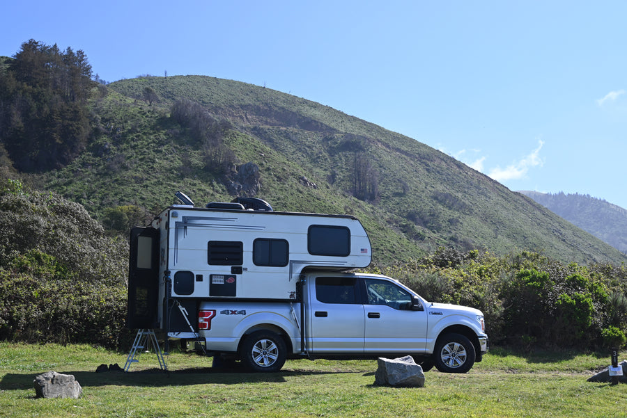 An RV rental truck camper on a silver pickup, parked on green grass with a large hillside behind it, ideal for camping.