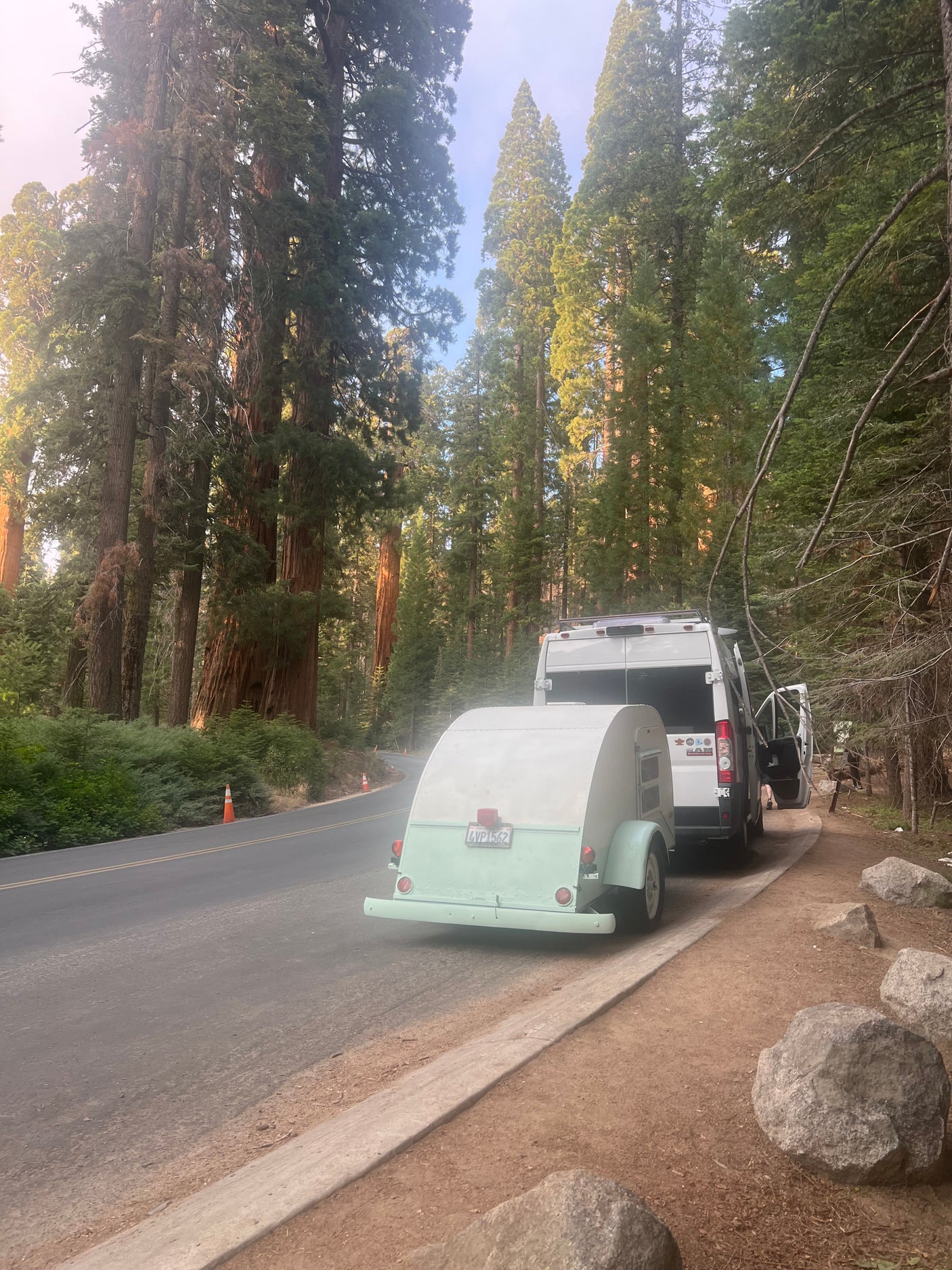 A white RV rental van tows a light green teardrop trailer alongside a road lined with towering redwood trees.