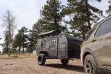 This RV rental features an off-road camper trailer and SUV parked among pine trees at a rustic campsite, ready for adventure.