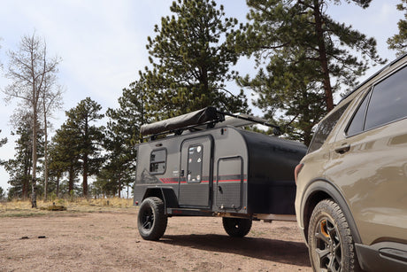 This RV rental features an off-road camper trailer and SUV parked among pine trees at a rustic campsite, ready for adventure.