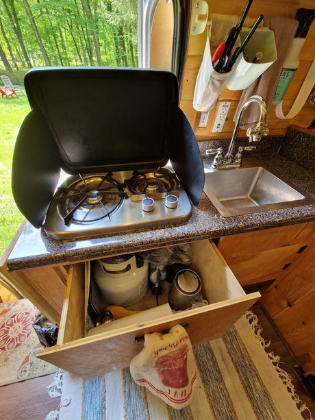 Inside an RV rental: A compact kitchen area featuring a two-burner stove, sink, and open drawer with propane tank.