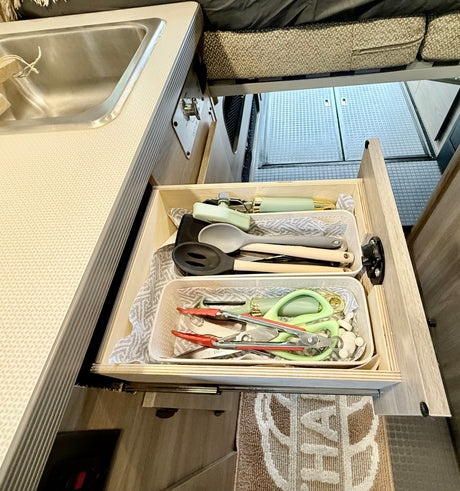 Open RV rental kitchen drawer with light green/grey utensils, next to a stainless steel sink and patterned counter.