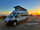 Experience an RV rental! Silver Ram ProMaster camper van with pop-top roof at sunset, parked overlooking mountains.