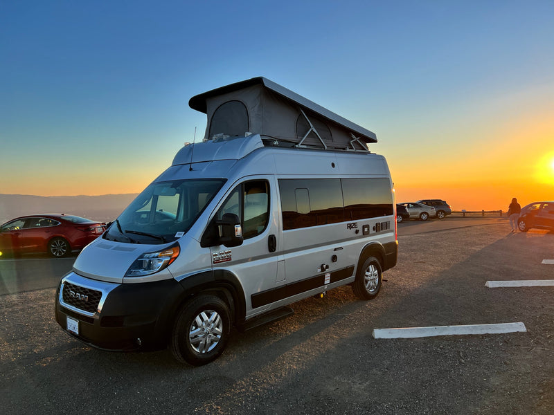  sits on a gravel lot under clear skies.|RV rental van at sunset. Wind turbines and a train define this desert road trip scene.|Silver RV rental camper van parked at sunset beside a long train & wind turbines.|Experience an RV rental! Silver Ram ProMaster camper van with pop-top roof at sunset — photo 5