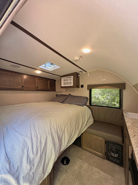 Cozy RV rental interior: a compact bedroom with striped bed, window AC, overhead storage, and floor vent.