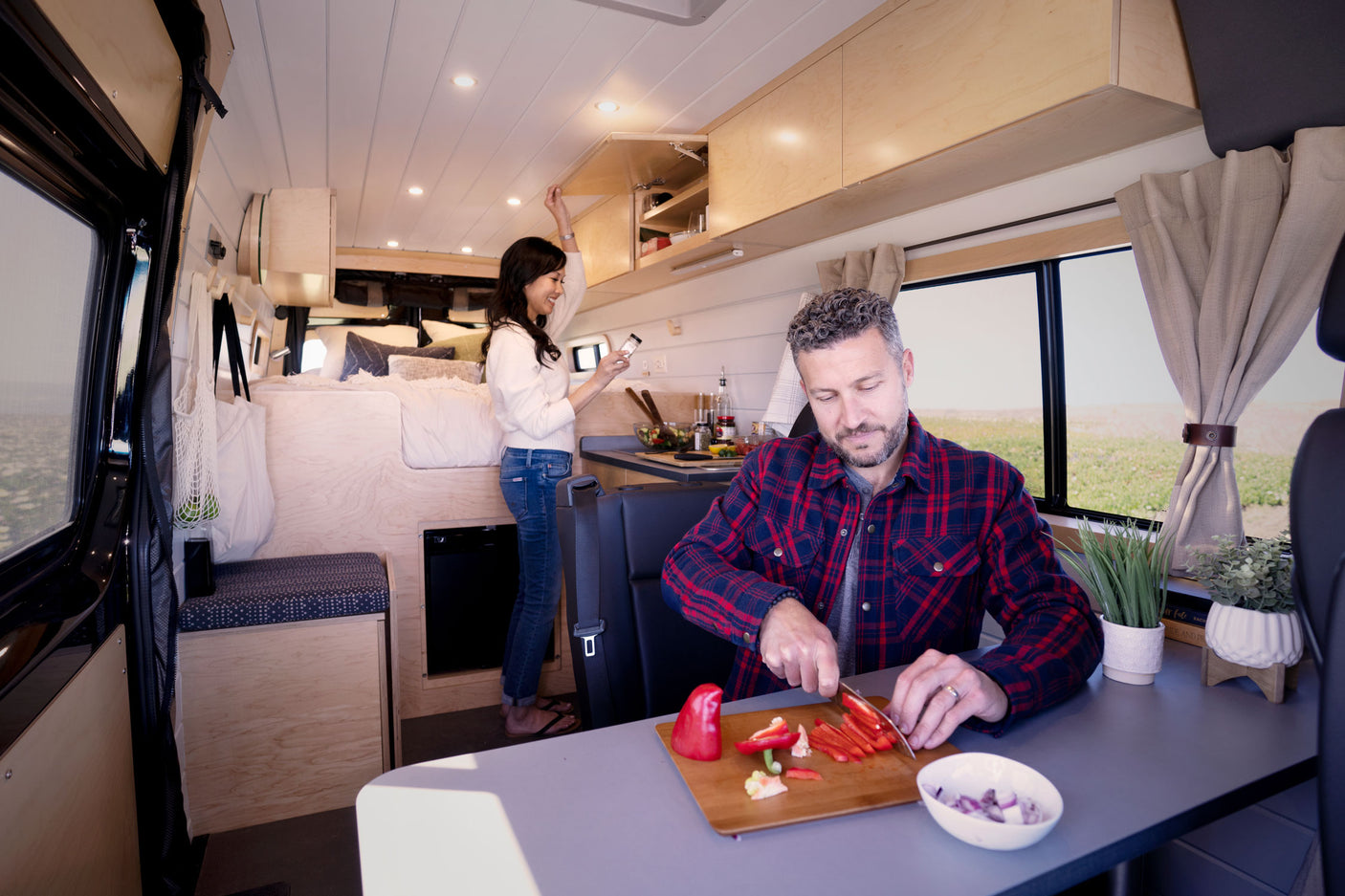 Couple prepares a meal in their modern RV rental. A man chops veggies while a woman accesses overhead storage in the cozy campervan.