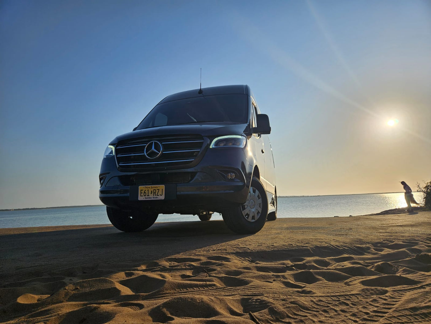 A dark Mercedes Sprinter **rv rental** sits on a sandy NJ beach at sunset, overlooking calm waters. Adventure awaits.