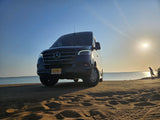 A dark Mercedes Sprinter **rv rental** sits on a sandy NJ beach at sunset, overlooking calm waters. Adventure awaits.