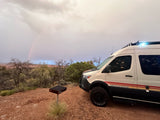 White Storyteller Overland RV rental parked at a scenic campsite. A faint rainbow arcs over a rugged landscape with a grill nearby.