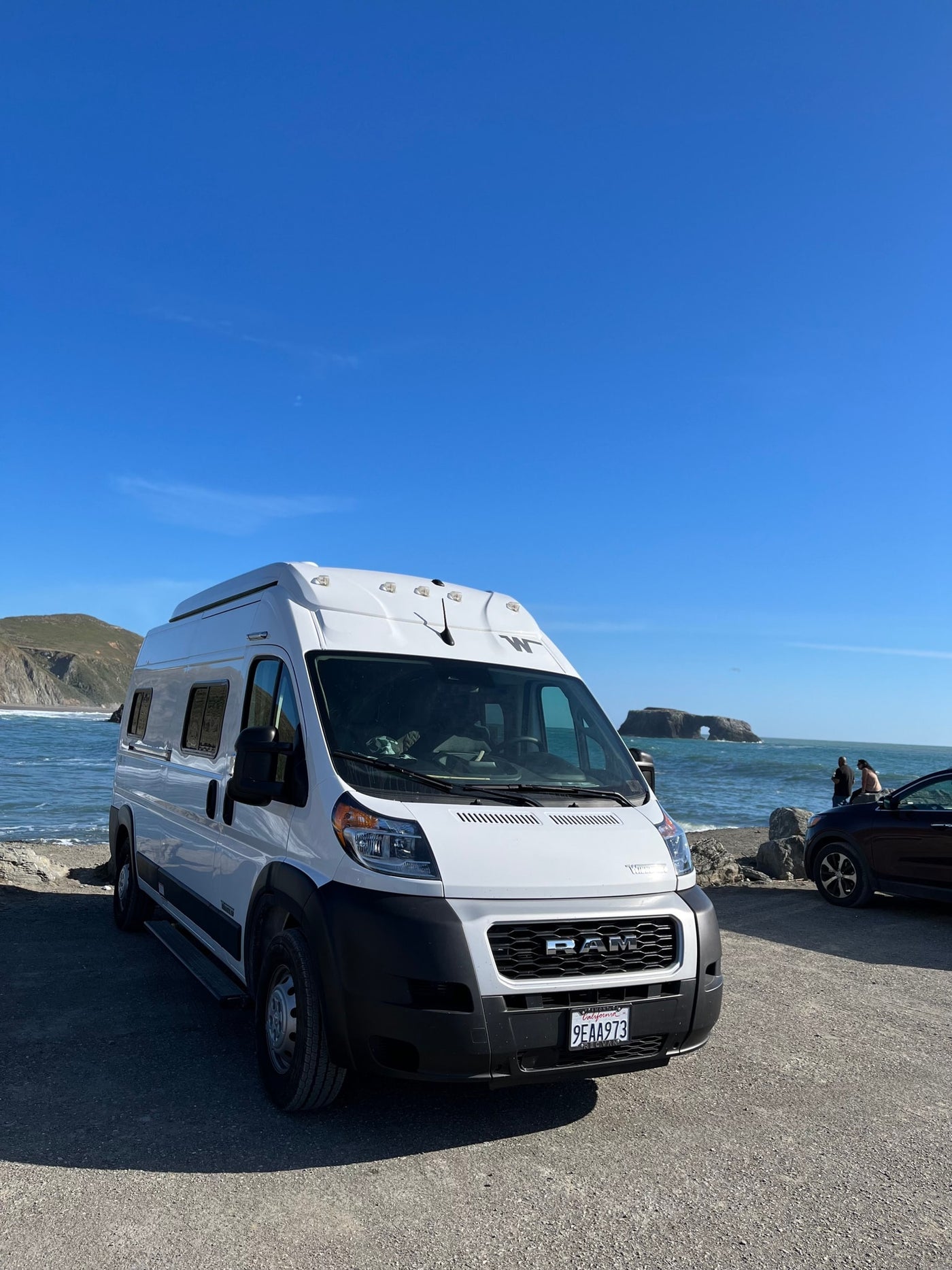 White RV rental parked at a sunny California coast, overlooking the ocean, a sea arch, and distant hills. — photo 3