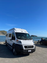 White RV rental parked at a sunny California coast, overlooking the ocean, a sea arch, and distant hills.