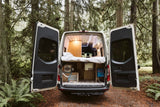 RV rental: Back of a custom conversion van in a forest, doors open showing a raised bed, utilities & storage.